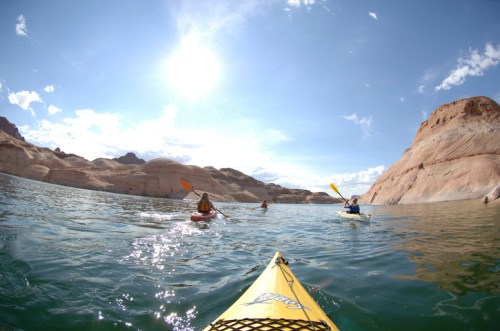 Two kayakers paddle on a calm lake surrounded by rocky cliffs under a bright blue sky. Sunlight reflects on the water.