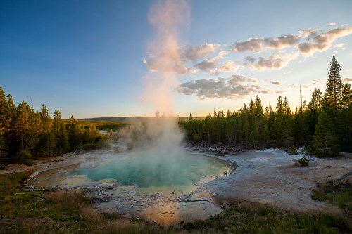 A steaming hot spring surrounded by trees at sunset, with a clear blue sky and wispy clouds in the background.