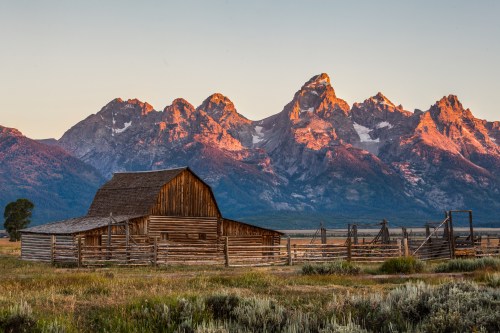 A rustic wooden barn in front of majestic mountains at sunrise, with a clear sky and lush greenery in the foreground.