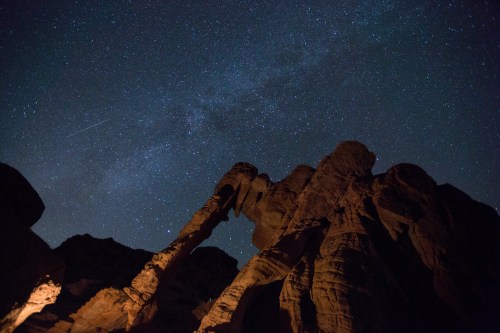 A starry night sky above towering rock formations, with a faint meteor streaking across the scene.