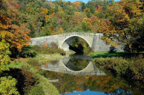A stone arch bridge spans a calm river, surrounded by vibrant autumn foliage reflecting in the water.