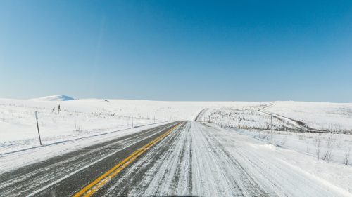 A snow-covered road stretches into the distance under a clear blue sky, surrounded by white fields.