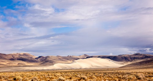 A vast desert landscape featuring rolling sand dunes under a cloudy sky, with mountains in the background.