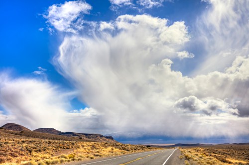 A wide, empty road stretches through a desert landscape under a dramatic sky filled with clouds.