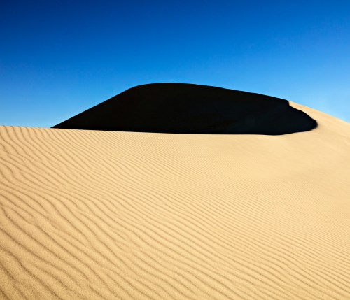 A smooth sand dune under a clear blue sky, with gentle ripples in the sand creating a serene landscape.