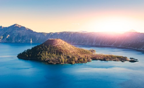 A serene lake with a small island surrounded by mountains, illuminated by a soft sunrise.