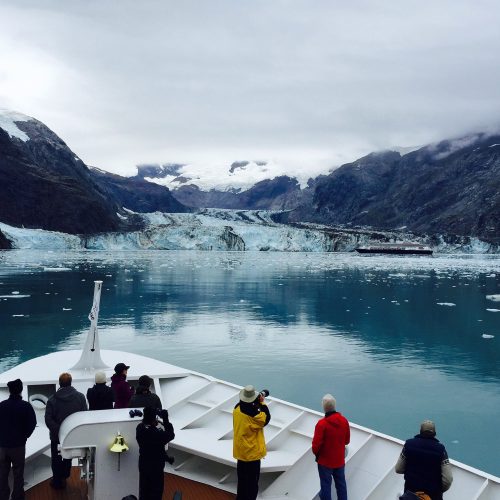 A group of people on a boat observing a glacier and icy waters surrounded by mountains under a cloudy sky.