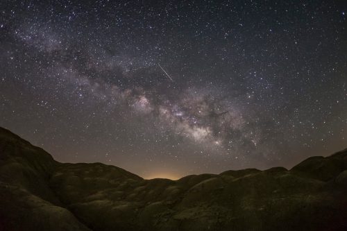 A stunning night sky filled with stars and the Milky Way, above dark, rolling hills. A shooting star is visible.