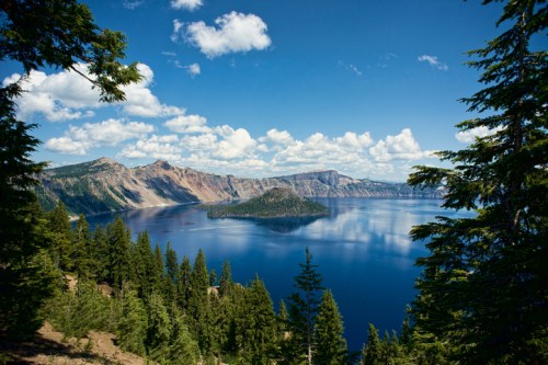 A serene view of Crater Lake surrounded by lush trees and mountains under a bright blue sky with fluffy clouds.