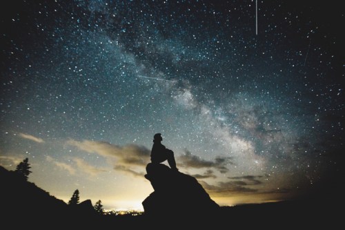 A silhouette of a person sitting on a rock, gazing at a starry sky filled with the Milky Way and shooting stars.
