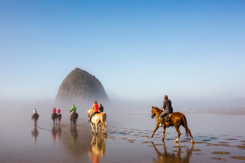A group of horseback riders strolls along a misty beach with a large rock formation in the background.