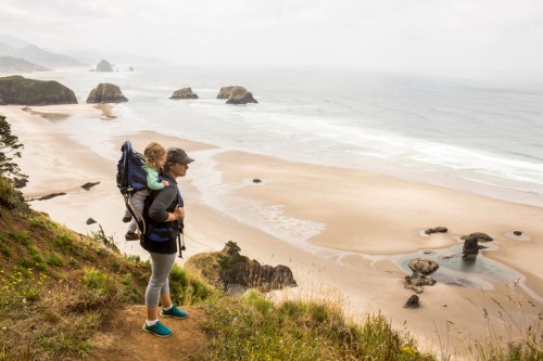 A woman with a child in a backpack stands on a cliff overlooking a beach and ocean, with rocky formations in the distance.