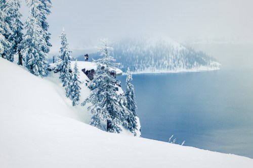 A snowy landscape with tall trees, a misty lake, and a person standing on a cliff overlooking the water.