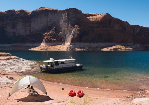 A serene lakeside scene with a houseboat, two kayaks, and people relaxing under a canopy on the sandy shore.