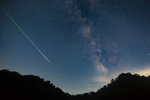 A starry night sky with the Milky Way and a bright meteor streaking across the scene above dark silhouettes of mountains.