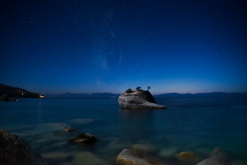 A serene night scene of a calm lake with a starry sky and a small island rock illuminated by starlight.