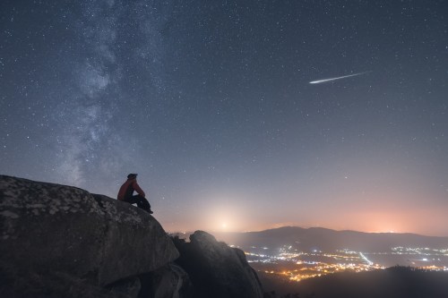 A person sits on a rock under a starry sky, gazing at the Milky Way and a shooting star above a distant city.
