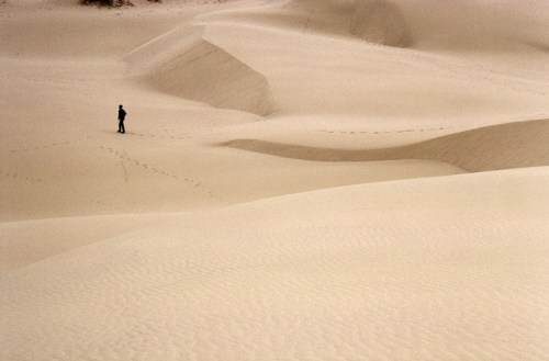 A solitary figure stands on vast, undulating sand dunes, with footprints trailing behind in the smooth, golden sand.