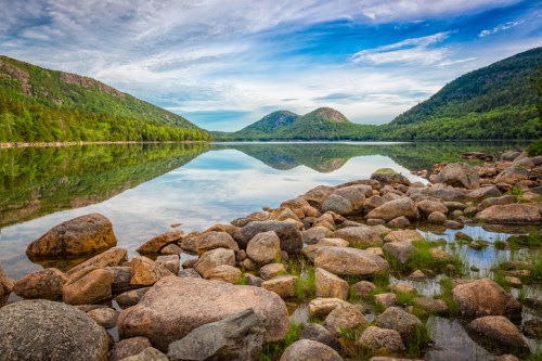 A serene lake surrounded by mountains, with a clear sky reflecting on the water and rocky shores in the foreground.