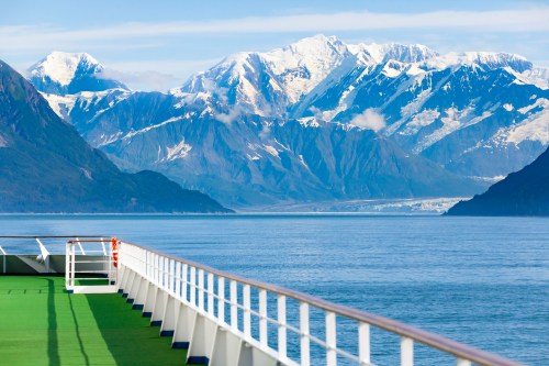 A ship's deck overlooks calm waters and majestic snow-capped mountains under a clear blue sky.