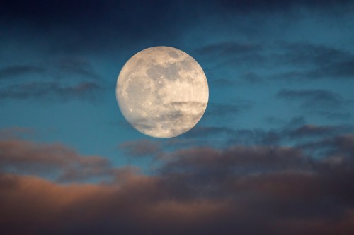 A bright full moon surrounded by wispy clouds against a blue sky at dusk.