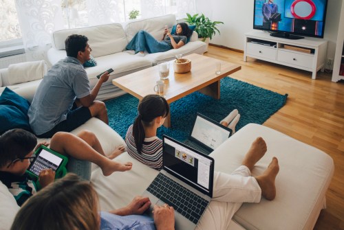A family relaxes in a living room, using devices while watching TV. A cozy atmosphere with a coffee table and blue rug.