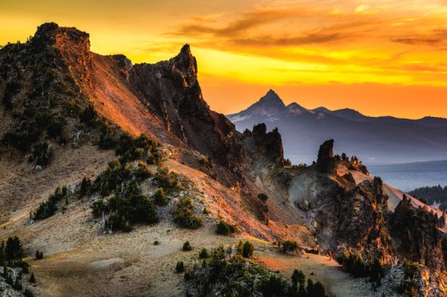 Sunset over rugged mountain peaks, with vibrant orange and yellow skies illuminating the landscape.