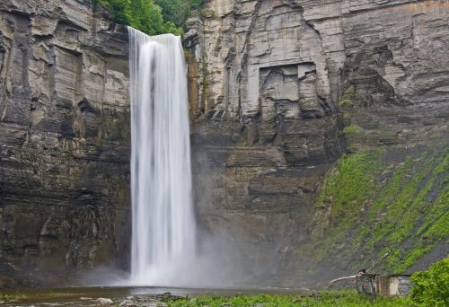 A tall waterfall cascades down rocky cliffs, surrounded by lush greenery and mist rising from the water below.