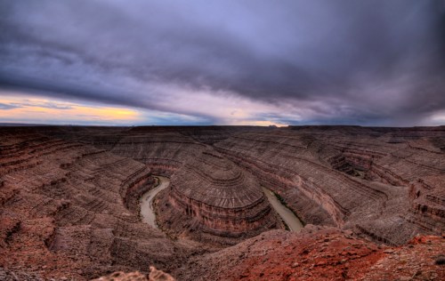 A dramatic canyon landscape with winding river, layered rock formations, and a cloudy sky at sunset.
