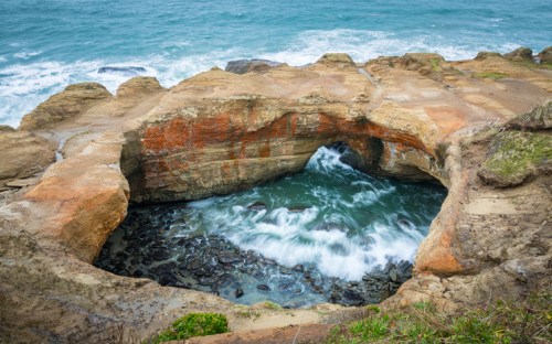 A rocky coastal formation with a circular opening, revealing turbulent water below and waves crashing against the shore.