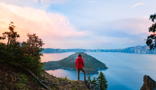 A person in a red jacket stands on a cliff, overlooking a serene lake surrounded by mountains and trees at sunset.