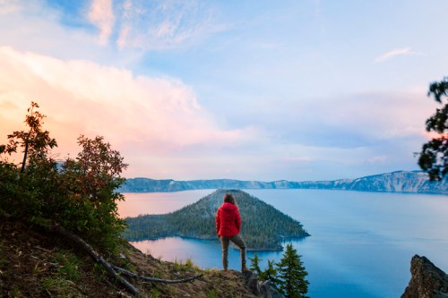 A person in a red jacket stands on a cliff, overlooking a serene lake surrounded by mountains and trees at sunset.