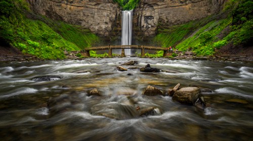 A scenic waterfall cascades down rocky cliffs, surrounded by lush greenery and a wooden bridge over a flowing river.
