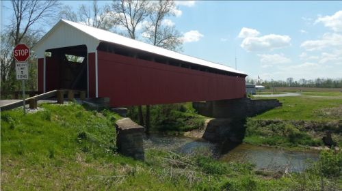 A red covered bridge spans a small creek, surrounded by green grass and trees under a blue sky with fluffy clouds.