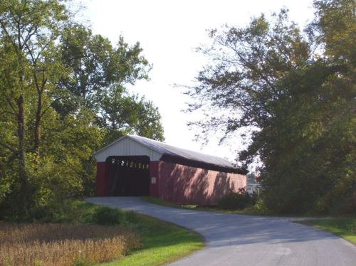 A red covered bridge surrounded by trees, with a winding road leading up to it on a sunny day.