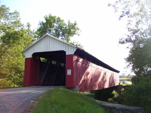 A red covered bridge with a white roof, surrounded by greenery, spanning a small creek on a sunny day.
