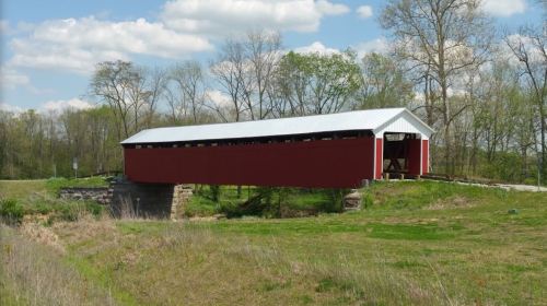 A red covered bridge spans a grassy area, surrounded by trees and a blue sky with fluffy clouds.