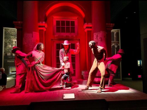 A dramatic red-lit scene depicting historical figures, including a woman, a child, and a man, in a tense moment.