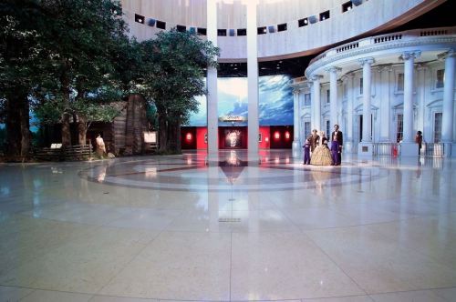 A spacious gallery with polished floors, large trees, and classical architecture, featuring visitors in elegant attire.