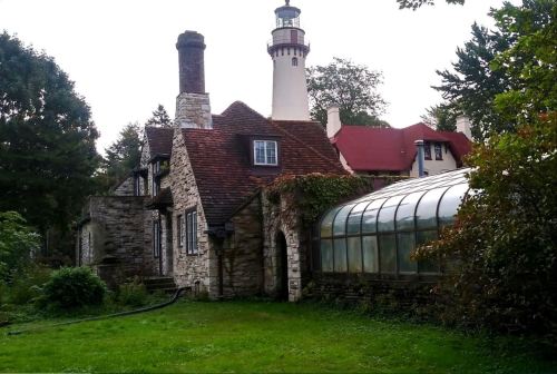 A stone house with a lighthouse tower and a glass greenhouse, surrounded by greenery and trees.