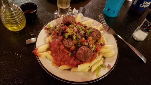 A plate of penne pasta topped with meatballs, marinara sauce, and a colorful vegetable mix, with drinks in the background.
