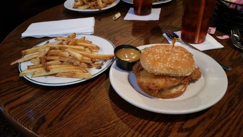 A plate with a sesame seed bun chicken sandwich and a side of crispy fries, accompanied by a small cup of dipping sauce.