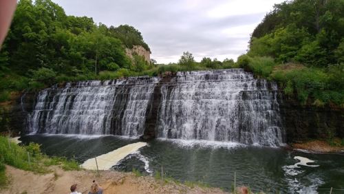 A scenic waterfall cascading over rocks into a calm pool, surrounded by lush greenery and a cloudy sky.