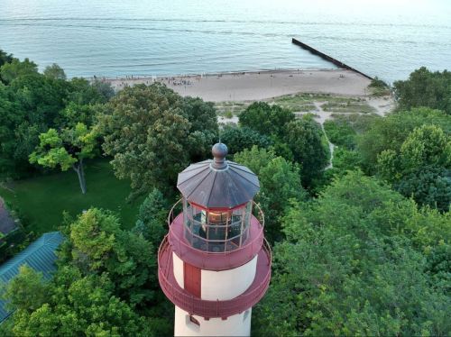 Aerial view of a lighthouse surrounded by trees, overlooking a sandy beach and calm water.