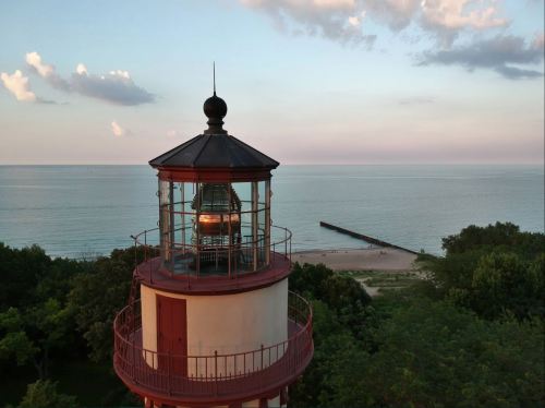 A lighthouse stands tall by the shore, overlooking a calm lake and sandy beach under a pastel sky.