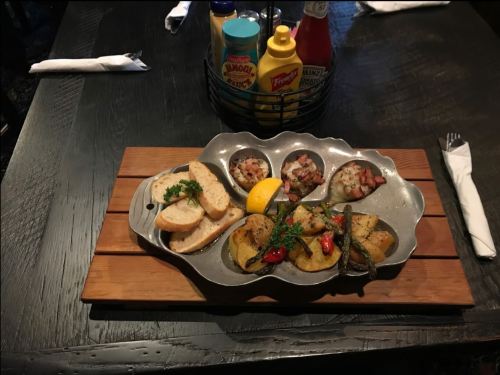 A metal platter with grilled seafood, lemon, and toasted bread, served on a wooden board with condiments in the background.