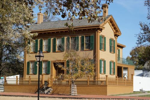 A historic two-story wooden house with green shutters and a porch, surrounded by trees and a lamp post.