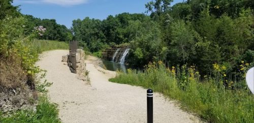 A scenic path leads to a waterfall surrounded by lush greenery and wildflowers under a clear blue sky.
