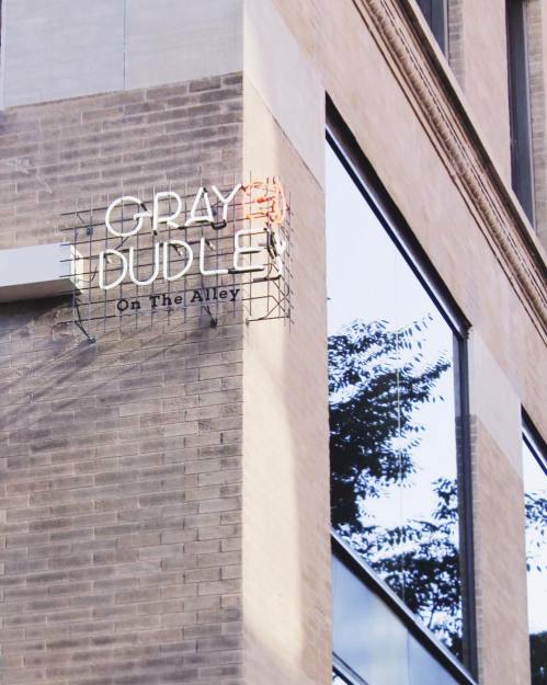 Sign for "Gray Dudes On The Alley" on a brick building, reflecting trees in nearby windows.