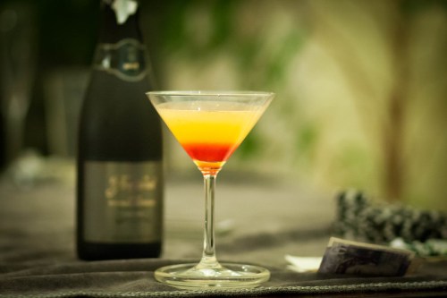 A colorful cocktail in a martini glass, with a bottle of sparkling wine in the background.